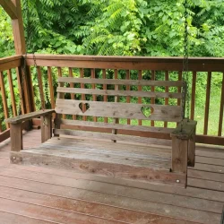 A wooden porch swing hangs from a chain on a covered deck, surrounded by lush green trees in the background.