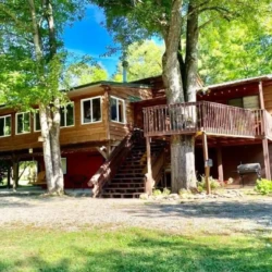 Wooden cabin with elevated deck, surrounded by trees, on a sunny day. Grass and gravel path in the foreground.