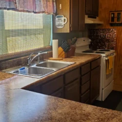 A kitchen with a dual sink, stove, and brown cabinets. A sign above the sink reads, "Never call me before coffee.