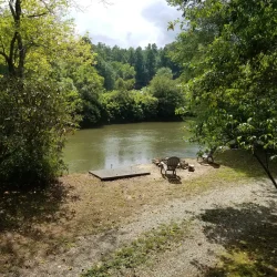 Riverside scene with a wooden chair on a small dock, surrounded by lush green trees and a gravel path in the foreground.