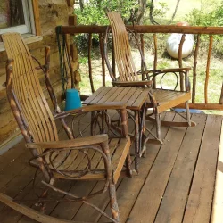 Two wooden rocking chairs on a rustic porch overlooking a grassy area, with sunlight casting shadows on the wooden floor.