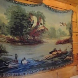 Tapestry depicting ducks flying and swimming in a natural landscape, hung on a wooden wall.