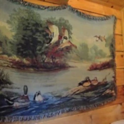 Tapestry depicting ducks flying and swimming in a natural landscape, hung on a wooden wall.