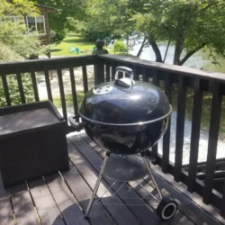 A black kettle grill on a wooden deck, overlooking a lawn and a body of water, surrounded by trees.