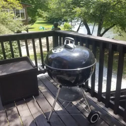 A black kettle grill on a wooden deck, overlooking a lawn and a body of water, surrounded by trees.