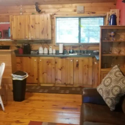 Cozy cabin kitchen with wooden cabinets, a round dining table, and a brown leather sofa. Pots hang above the counter.