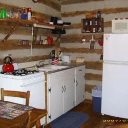 Rustic kitchen with white appliances, wooden shelves, and log walls. Includes a stove, fridge, microwave, and a small dining table.