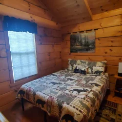 A small wooden cabin bedroom with a double bed, bear-themed bedding, a window with a sheer curtain, and rustic wall art.