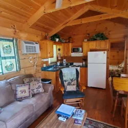 Cozy wooden cabin interior with a ceiling fan, couch, coffee table, kitchen area, and dining table. Air conditioner on the wall.