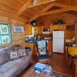 Cozy wooden cabin interior with a ceiling fan, couch, coffee table, kitchen area, and dining table. Air conditioner on the wall.