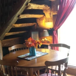 Cozy wooden dining area with a round table, four chairs, floral centerpiece, and stairs in a rustic setting.
