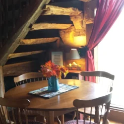 Cozy wooden dining area with a round table, four chairs, floral centerpiece, and stairs in a rustic setting.