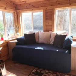 A sunlit room with a blue sofa, multiple cushions, large windows, a wooden interior, and a wood-burning stove on the left.