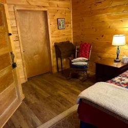 A cozy wooden room with a bed, quilt, rocking chair, desk, and lamp. Closed wooden door on the left. Warm lighting creates a homey feel.