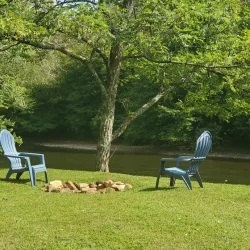 Two blue chairs face each other under a tree beside a river, with a circle of stones on grassy ground.