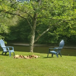 Two blue chairs face each other under a tree beside a river, with a circle of stones on grassy ground.