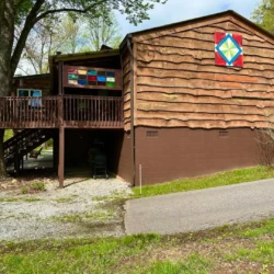 Wooden house with a colorful quilt pattern on the side, surrounded by trees and a driveway in front.