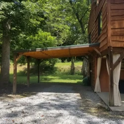 Wooden carport attached to a rustic house, set against trees and grass, with gravel driveway in the foreground.