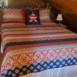 Colorful bed with patterned quilt and star pillow in a wooden room, featuring a small lamp and window air conditioner.