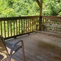 Wooden deck with a bench and chair, surrounded by green trees and sunlight filtering through.