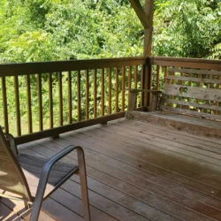 Wooden deck with a bench and chair, surrounded by green trees and sunlight filtering through.