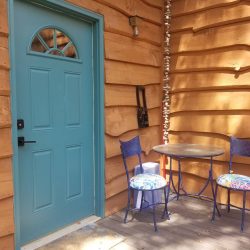 Blue door and two blue chairs with colorful cushions on a porch. Round table and wooden wall with wavy texture in the background.
