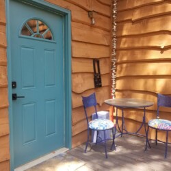 Blue door and two blue chairs with colorful cushions on a porch. Round table and wooden wall with wavy texture in the background.