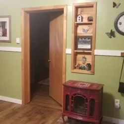 Room with green walls, a wall clock, open door, shelves with decorations, and a red wood stove on a wooden floor.