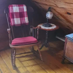 Cozy attic corner with a wooden rocking chair, red seat cushion, plaid blanket, small side table, and antique lamp.