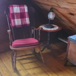 Cozy attic corner with a wooden rocking chair, red seat cushion, plaid blanket, small side table, and antique lamp.