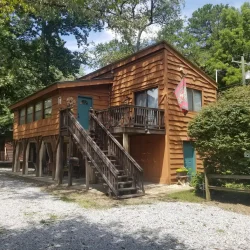 Two-story wooden cabin with a rustic exterior, staircase leading to the upper floor, and surrounded by trees on a gravel path.