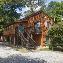 Two-story wooden cabin with a rustic exterior, staircase leading to the upper floor, and surrounded by trees on a gravel path.
