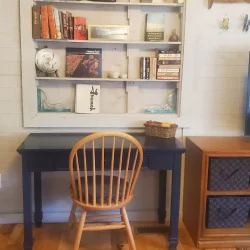 Wooden chair at a black desk with a bookshelf above, decorated with books and decorative items. Nearby TV on a stand.