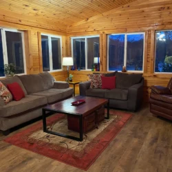 A cozy living room with wooden walls, two sofas, an armchair, a coffee table, and decorative pillows and lamps.