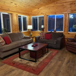 A cozy living room with wooden walls, two sofas, an armchair, a coffee table, and decorative pillows and lamps.
