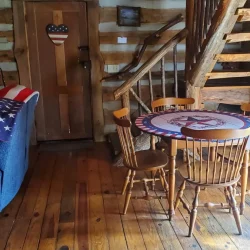 Rustic interior with wooden floors, a log wall, a small round table with chairs, a staircase, and a cabinet with a patriotic theme.