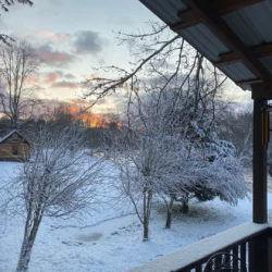 Snow-covered backyard with trees at sunset, viewed from a porch with a wind chime and a shed in the distance.