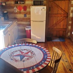 Rustic kitchen with a wooden table and two chairs, fridge, microwave, and a door. Chandelier above, exposed beams visible.