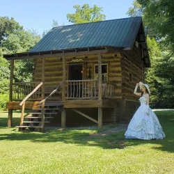 A woman in a long dress and hat stands on grass in front of a small wooden cabin surrounded by trees.