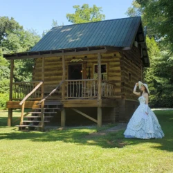 A woman in a long dress and hat stands on grass in front of a small wooden cabin surrounded by trees.