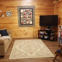 Cozy room with wooden walls, a sofa, TV, quilt on the wall, and a rocking chair. Bookshelf filled with games and books.