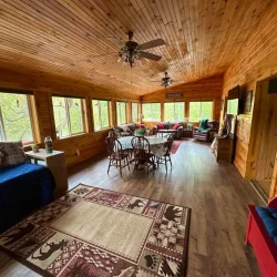 Rustic sunroom with wooden walls, ceiling fans, large windows, sofas, and a dining table with chairs on wood flooring.