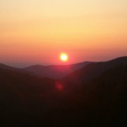Sunset over mountain range with a clear sky and the sun near the horizon, casting orange and pink hues across the landscape.
