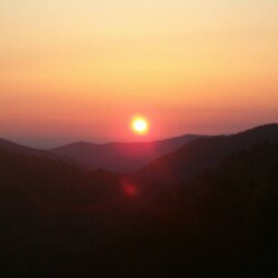 Sunset over mountain range with a clear sky and the sun near the horizon, casting orange and pink hues across the landscape.