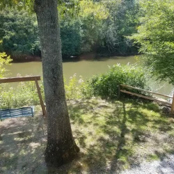 A wooden swing facing a river surrounded by trees. A tree and a wooden fence are in the foreground.