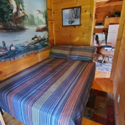 Cozy cabin bedroom with a striped bedspread, wooden walls, and wildlife-themed artwork. Doorway leads to a kitchen area.