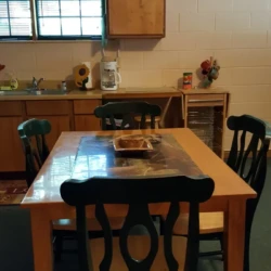 A kitchen with a wooden table and four chairs, cabinets, a window with green curtains, a clock, and various items on the counter.