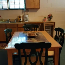 A kitchen with a wooden table and four chairs, cabinets, a window with green curtains, a clock, and various items on the counter.