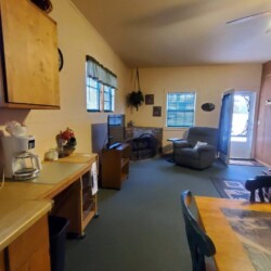 Cozy living area with a recliner, TV, wooden cabinets, and a dining table. Windows and a door are visible in the background.