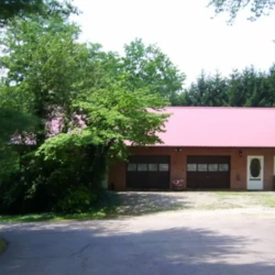 A two-door brick garage with a red roof surrounded by trees and bushes on a sunny day.